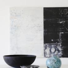 Modern interior design image of Bernd Haussmann artwork featuring home decor in shades of black, brown and teal.  There is a wood bowl, an orthoceras bowl and a teak sphere on a white table. 