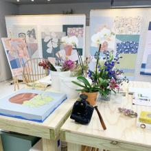 this is the studio of taelor fisher with paintings on a moveable wall and flowers and paints on a table. 